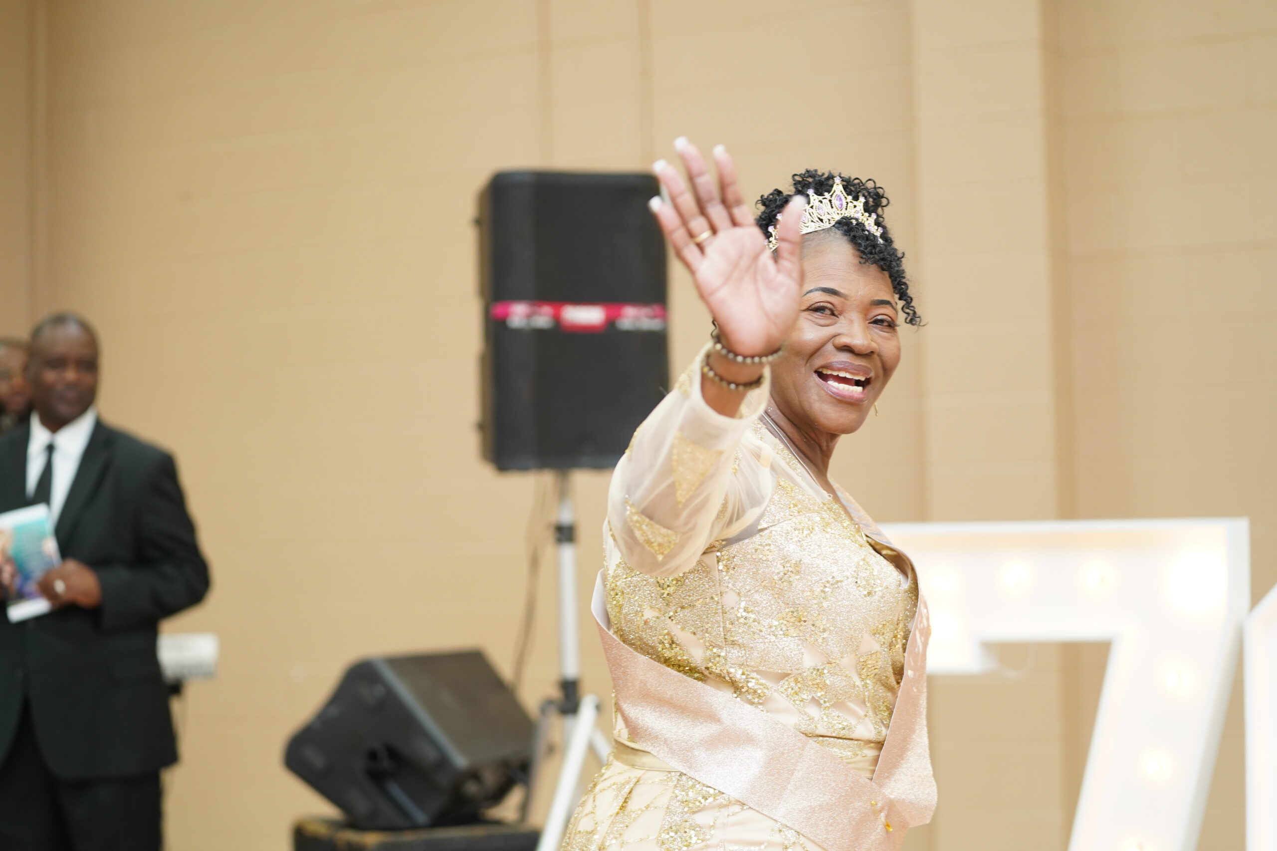 Elizabeth Hemingway Bowen smiling and waving at her 70th birthday celebration, wearing a golden gown and a tiara, with a joyful expression in a festive setting.